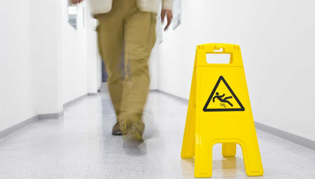 Person walking past a yellow wet floor sign in a white hallway, cautioning against slip hazards