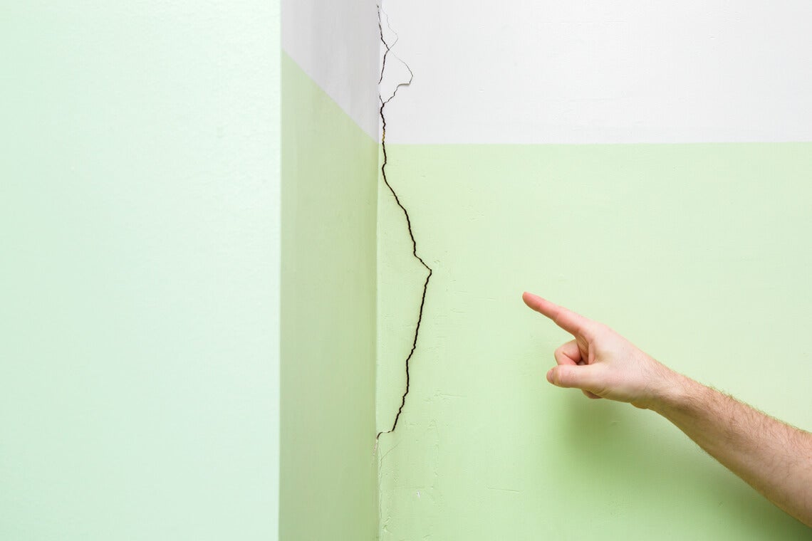 Man's hand finger pointing to cracked corner wall in house. Building problems and solutions concept.