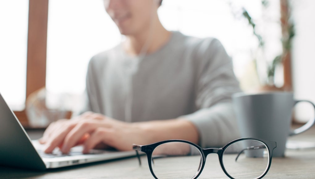 Cut view of a guy sitting at the table and wokring. He is wokring using the laptop. Besides the laptop there is a cup of tea. There are glasses in the front of the picture