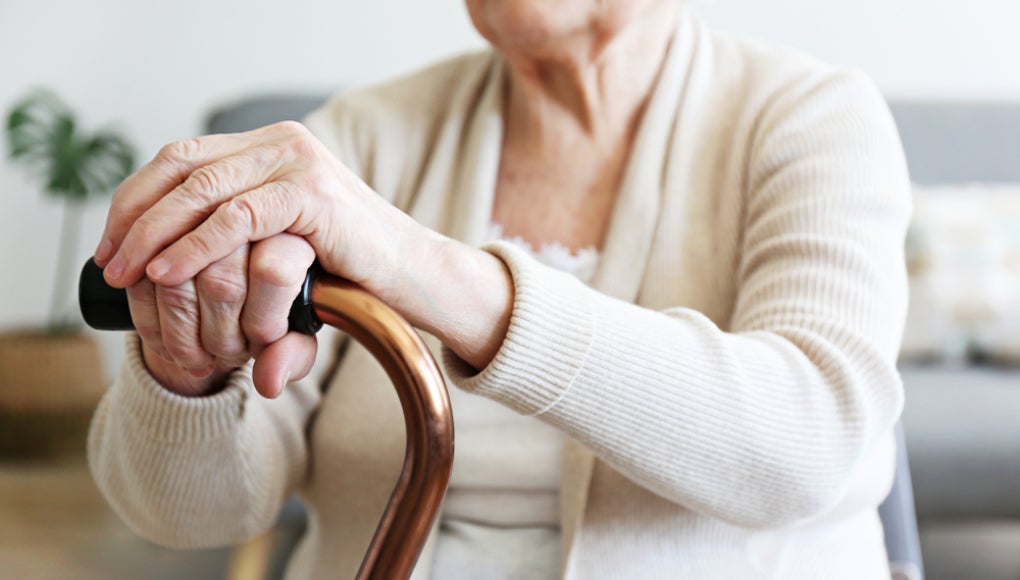 Elderly woman sitting in nursing home room holding walking quad cane with wrinked hand. Old age senior lady wearing beige cardigan, metal aid stick handle bar close up.
