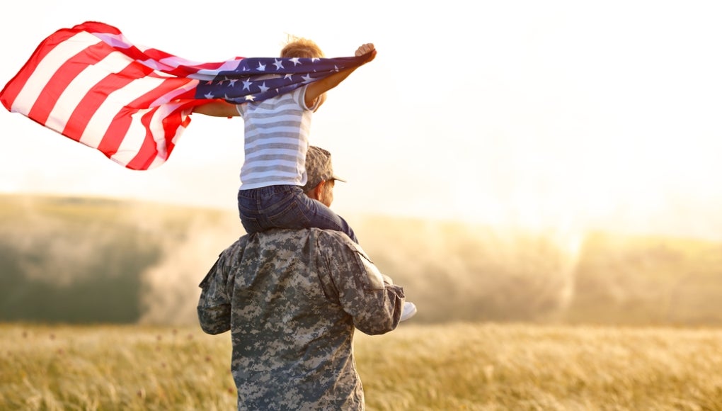 Rear view of military man father carrying happy little son with american flag on shoulders and enjoying amazing summer nature view on sunny day, happy male soldier dad reunited with son after US army