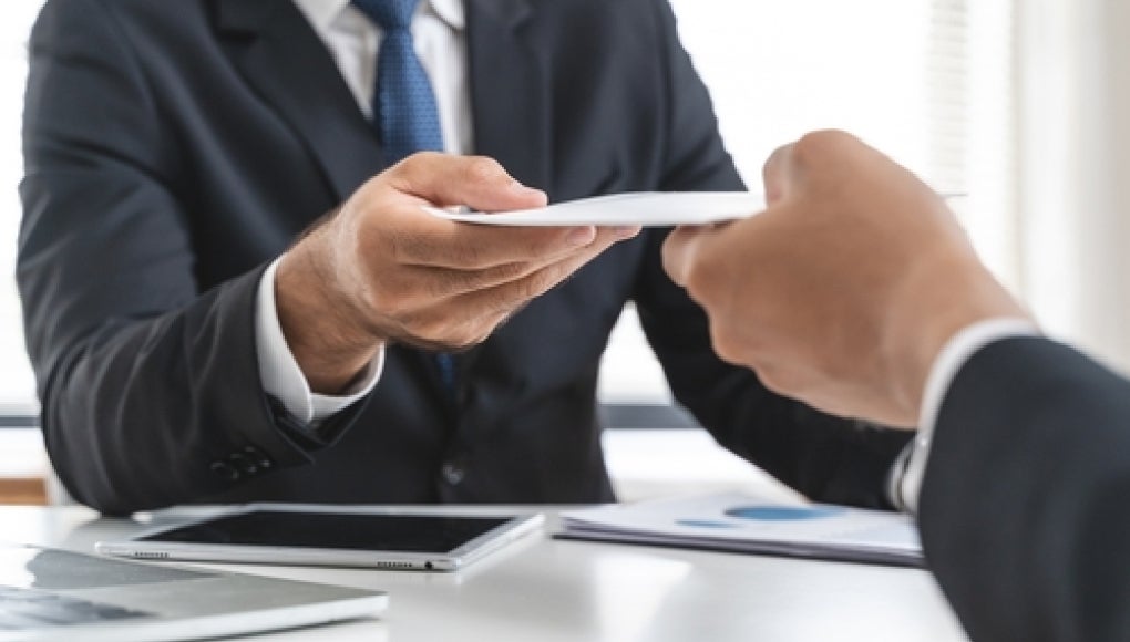 two suited employees exchanging notes, a tablet between them 