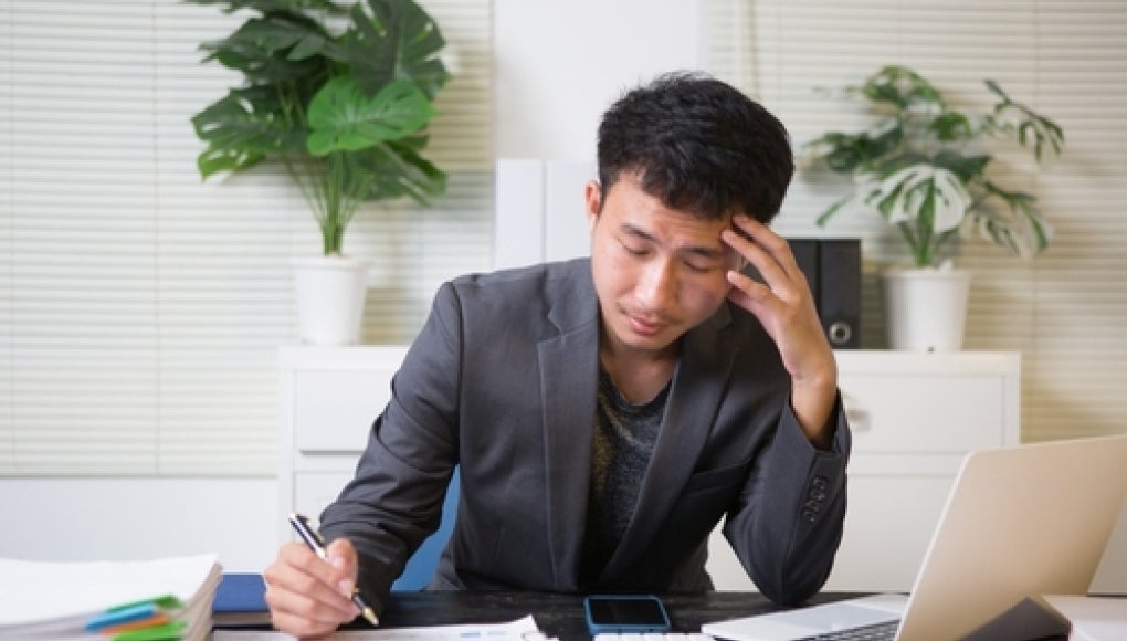 person sitting at desk with piles of papers, laptop nearby 
