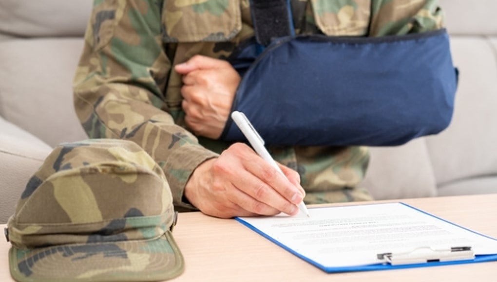veteran with arm in cast sitting on couch filling out a form on a clipboard with a pen in hand.