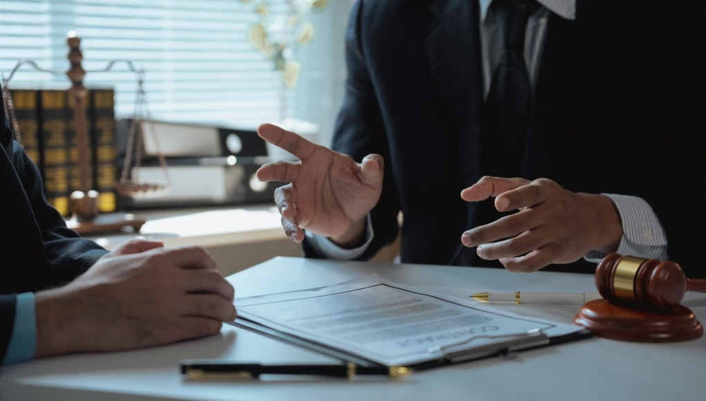 Lawyer gesturing with hands while discussing contract details with client in office, surrounded by legal documents and a gavel resting on the desk, facilitating clear communication