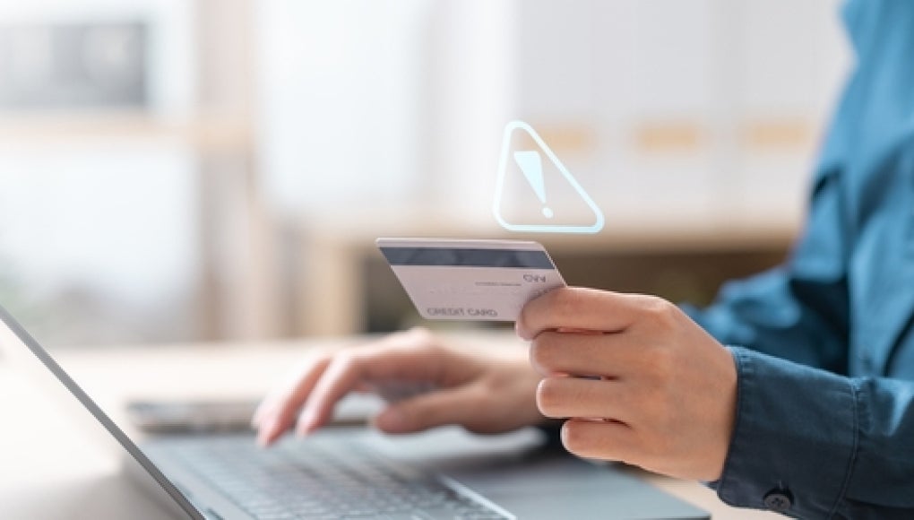 person sitting in front of laptop holding a bank card with a error sign indicating fraud
