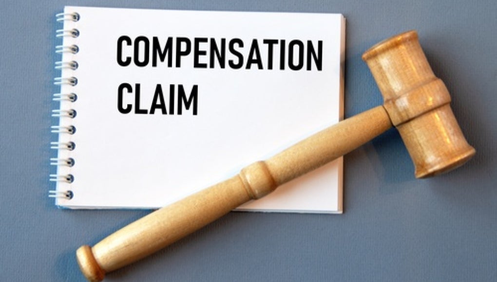 notepad with the words 'compensation claim' on it, with a grey background and wooden gavel resting on top of it 