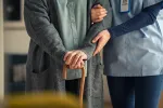 Close up hands of caregiver doctor helping old woman at clinic. Nurse holding a senior patient with walking stick. Elder woman using walking cane at nursing home with nurse holding hand for support. 