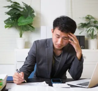 person sitting at desk with piles of papers, laptop nearby 