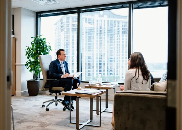 Man in business attire sitting in a modern office, speaking with two colleagues facing him. Large windows overlook tall buildings, creating a professional atmosphere.