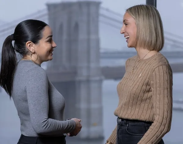 Two people stand in conversation, smiling at each other. They are in front of a large window with the Brooklyn Bridge visible in the background.