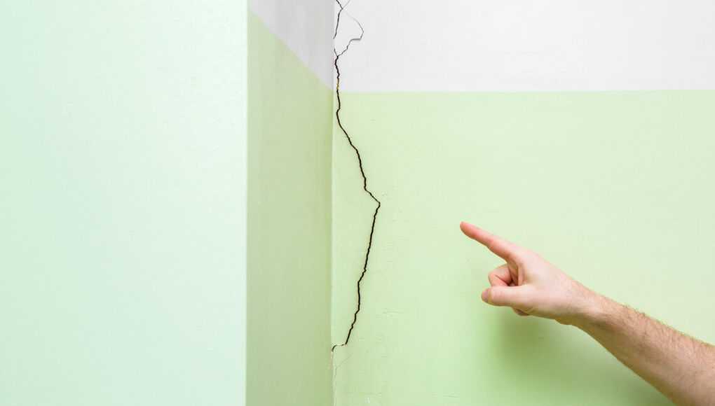 Man's hand finger pointing to cracked corner wall in house. Building problems and solutions concept.