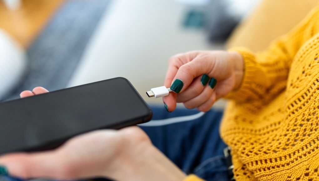 Woman in yellow sweater plugging USB cable into smartphone, close-up of hands and device on couch background.
