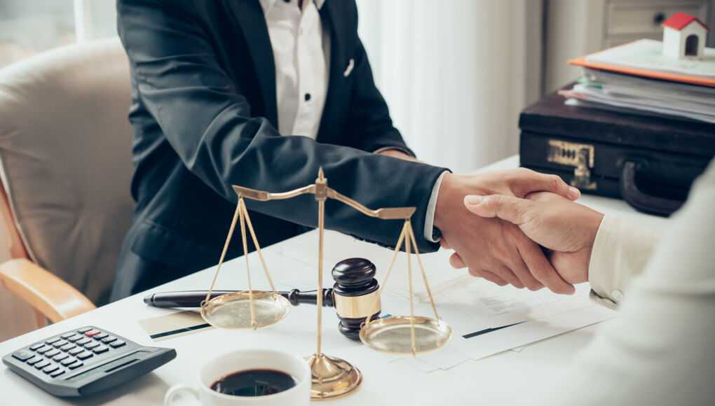 Lawyer shaking hands with client in office with legal balance scale and gavel on desk.