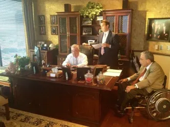Three men are in a well-furnished office. One sits at a large wooden desk using a computer, another stands with papers, and the third sits in a wheelchair. The mood is professional.