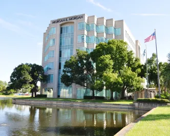 A modern office building with reflective glass windows is situated beside a serene pond. Two large trees and flagpoles with U.S. and company flags stand nearby.