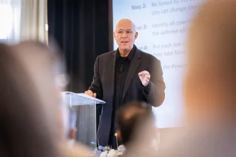 A man in a dark suit speaks confidently at a podium, with a projected presentation behind him.