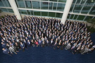 A large group of people in formal attire stands closely together in a bright, spacious indoor area with large windows, conveying a sense of unity and professionalism.